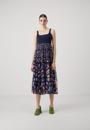 Abito floreale blu navy con corpetto aderente e gonna a strati in chiffon trasparente caratterizzata da un motivo di fiori colorati. Scarpe con tacco verde completano l'outfit.