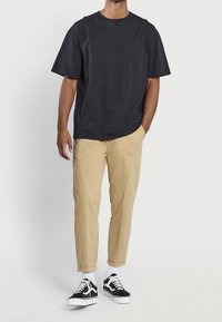 T-shirt noir en coton à manches courtes, pantalon beige fuselé et sneakers noires. Détail avec lacets blancs et chaussettes de cheville visibles.