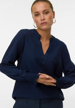 Vrouw in een marineblauwe blouse met lange mouwen, met gathered manchetten en een split halslijn, poseert met de handen samengevouwen voor een effen achtergrond.
