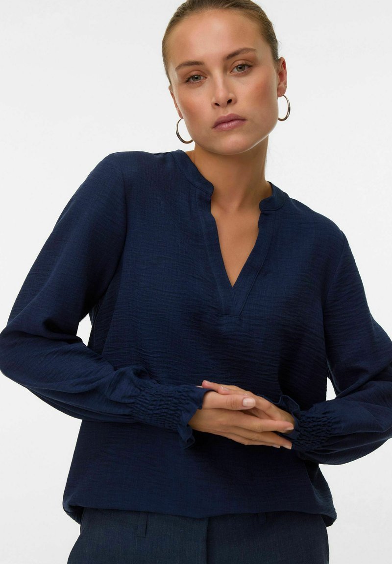 Donna che indossa una blusa a maniche lunghe blu navy con polsini arricciati e scollatura profonda, posando con le mani giunte davanti a uno sfondo semplice.