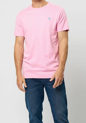 Mann trägt ein schlichtes rosa T-Shirt mit kurzen Ärmeln, einem kleinen blauen Logo auf der Brust und dunkelblauen Jeans vor schlichtem Hintergrund.