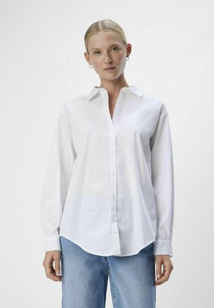 White button-down shirt made of cotton, featuring long sleeves, a classic collar, and a curved hem, paired with light blue jeans.
