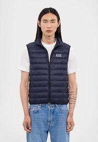 Jūras spēku puffer veste ar augstu apkakli, quilted struktūru, priekšējo rāvējslēdzēju un izšūtu logo; valkāta virs baltas krekliņas un ziliem džinsiem.