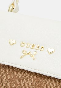 Borsa sintetica bianca con hardware dorato, logo "GUESS Girl" e dettagli a forma di cuore; sfondo marrone con motivo e finitura testurizzata.