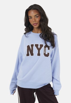 Vrouw met lang donker haar die een lichtblauwe trui draagt met "NYC Athletic" in grote letters en een zwarte broek met witte strepen.