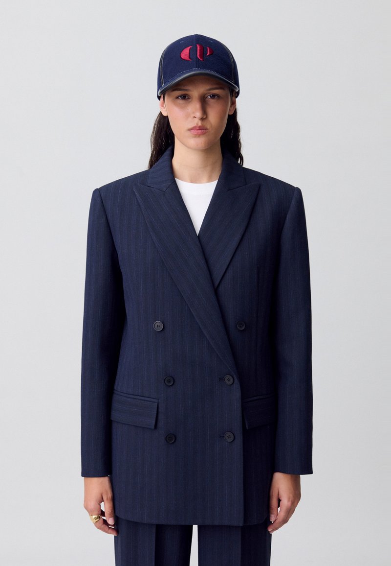 Blazer de doble botonadura a rayas marineras con grandes solapas, dos botones y bolsillos laterales; combinado con una gorra azul marino que presenta un logo rojo.