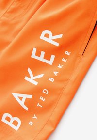 Svijetlo narančasta tkanina s bijelim tekstom "BAKER by Ted Baker" ispisanim dijagonalno blizu okomite šavove i bočnog džepa.