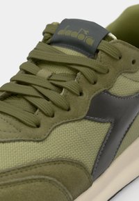 Green athletic shoe featuring mesh and suede materials, rounded toe, flat laces, and a dark gray logo patch on the tongue.