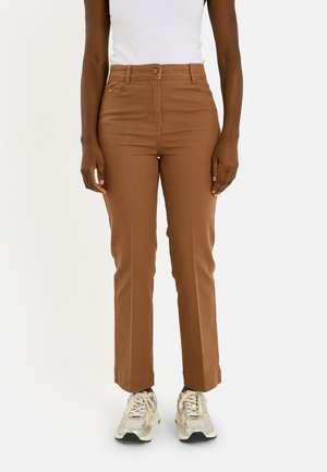 Personne portant un pantalon marron taille haute et des baskets blanches, associée à un haut blanc sans manches rentré, debout devant un fond uni.