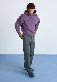 Junge Person in einem lila Hoodie mit dem Text "Alpha Industries", grauen Cargo-Hosen und lila Turnschuhen, die auf einem blauen Boden steht.