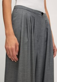 Pantalons plissés gris avec une fermeture à bouton, confectionnés en tissu texturé. Présentent une taille ajustée et une coupe décontractée.