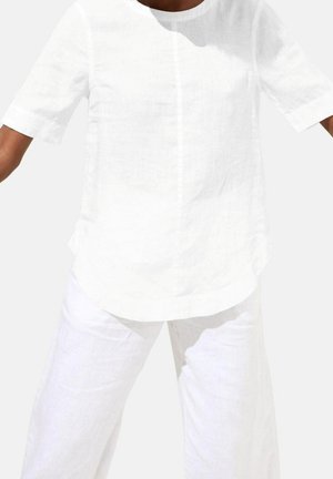 Personne portant une chemise large en lin blanc à manches courtes et un pantalon large assorti, debout avec les bras légèrement écartés.