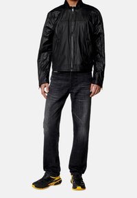 Chaqueta negra ligera con superficie texturizada, cierre de cremallera y bolsillos laterales, combinada con jeans de mezclilla oscuros y zapatos negros con detalles amarillos.