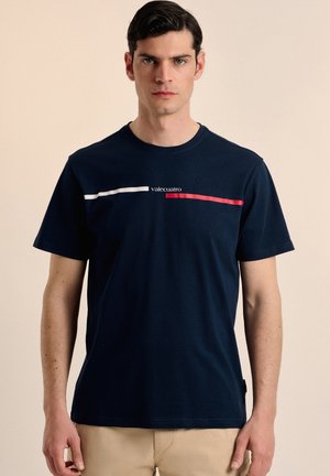 Camiseta de algodón azul marino con un diseño de rayas blancas y rojas en el pecho, con el texto "valecuatro" en el centro. Mangas cortas.