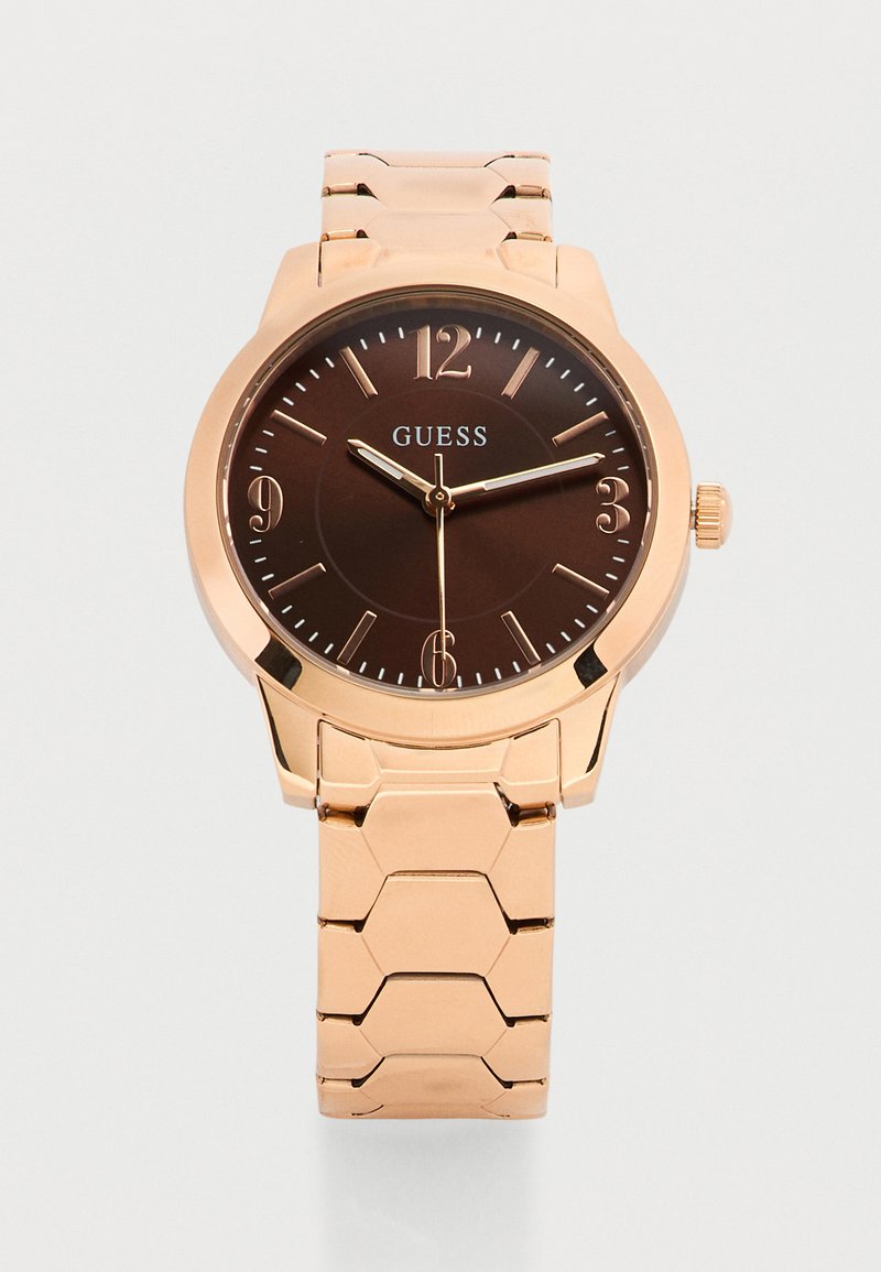 Ρολόι χειρός από ροζ χρυσό μέταλλο με καντράν σκούρου καφέ, χρυσούς δείκτες ώρας, τρία δείκτες και το λογότυπο GUESS στο καντράν.