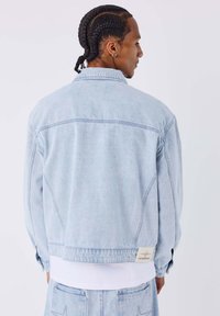 Giacca in denim azzurro chiaro con vestibilità ampia, dotata di colletto, maniche lunghe e una toppa con logo bianco sull'orlo posteriore.