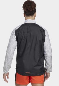 Leichte Jacke mit einem schwarzen Rücken, gemusterten grauen Ärmeln und einem reflektierenden Design. Verfügt über elastische Bündchen und einen abgerundeten Saum.