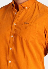 Oranje corduroy shirt met een button-down kraag, enkele borstzak en contrasterende witte knopen. Heeft een klein zwart logo op de zak.