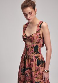 Blumenkleid in Pink, Braun und Schwarz, mit einer Schleifendetails am Ausschnitt und ausgeschnittenen Seiten. Das Material wirkt glatt und leicht.