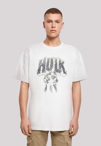 Hvit bomulls t-skjorte med et grafisk motiv av en knyttet neve og "HULK"-tekst i grått med grønne detaljer. Korte ermer og avslappet passform.