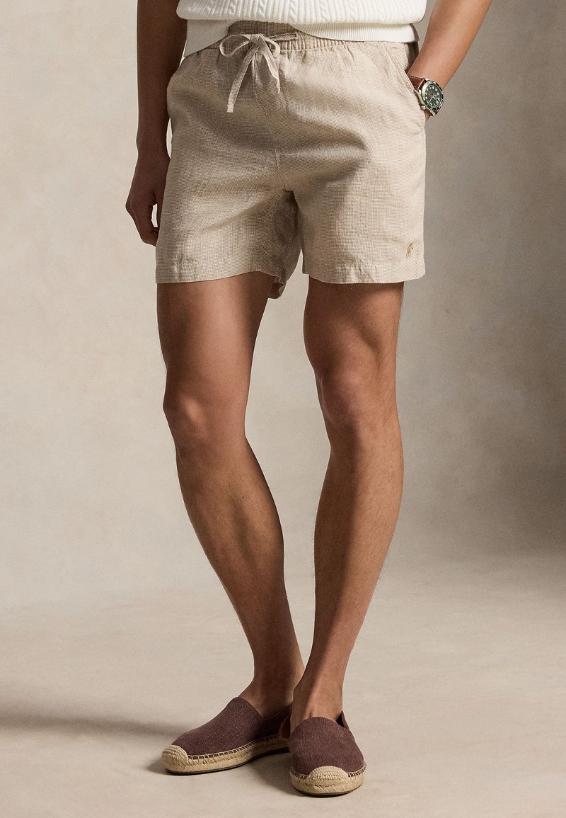 Μπεζ σορτς με κορδόνι που φοριέται με καφέ slip-on εσπαντρίγιες, λευκό πλεκτό τοπ και ένα ρολόι στον αριστερό καρπό.