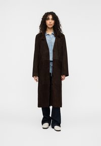 Καφέ σουέντ μακρύ παλτό με χαλαρή εφαρμογή, δύο μπροστινές τσέπες και κουμπιά. Φορεμένο πάνω από ανοιχτό μπλε πουκάμισο με γιακά και σκούρα τζιν με καμπάνα.