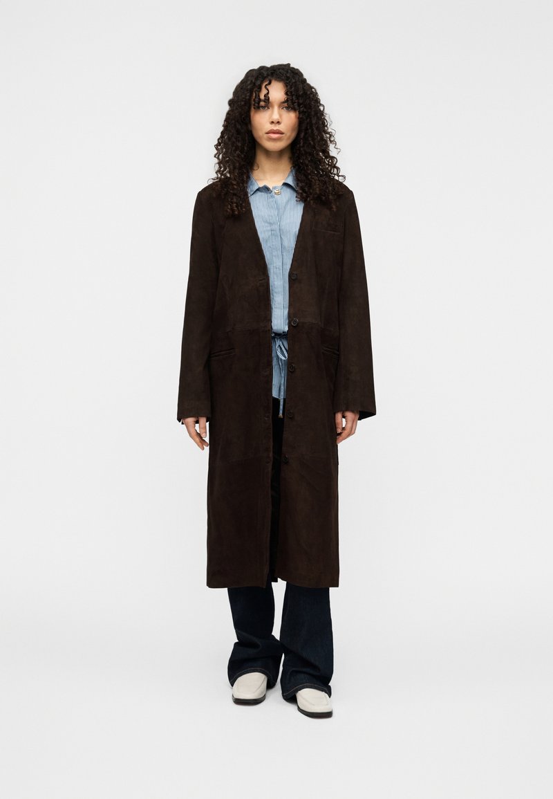 Καφέ σουέντ μακρύ παλτό με χαλαρή εφαρμογή, δύο μπροστινές τσέπες και κουμπιά. Φορεμένο πάνω από ανοιχτό μπλε πουκάμισο με γιακά και σκούρα τζιν με καμπάνα.