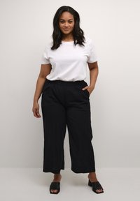 Pantaloni neri a gamba larga con elastico in vita, abbinati a una camicia bianca a maniche corte. Il modello sta in piedi con sicurezza, indossando sandali neri aperti.