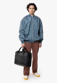 Sininen kevyt bomber-takki, musta suorakulmainen laukku, jossa on yläkahvat, ruskeat housut ja valkoiset kengät, joissa on keltaiset pohjat.