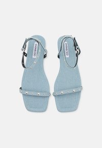 Sandalias de mezclilla azul con dos tiras estrechas, pequeños tachones plateados, correa en el tobillo, suela plana y un diseño sencillo y moderno.