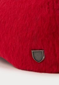 Rode fuzzy stof met een klein metalen schildvormig embleem met het label "RAYMON" nabij de rand op een witte achtergrond.