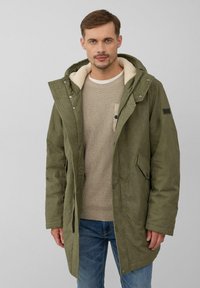 Olijfgroen parka met een zachte, crèmekleurige fleece voering, ritssluiting en drukknopen, zijzakken en een capuchon. Textuurdetails van de stof zichtbaar.