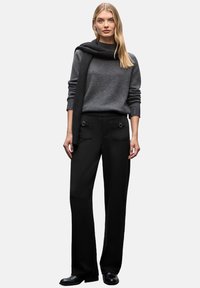 Maglione grigio a maglia, drappeggiato con una sciarpa nera, abbinato a pantaloni neri a vita alta con tasche frontali, tenuti da dettagli metallici.