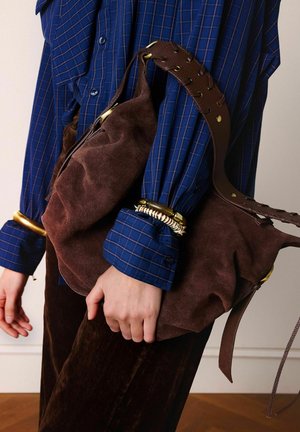 Persona che indossa una camicia blu a quadri e pantaloni marroni, che tiene una grande borsa a tracolla in suede marrone con una larga tracolla in pelle e braccialetti dorati.