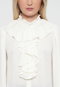 White blouse with a ruffled collar and front detail, featuring a smooth texture and button closure, designed with long sleeves.