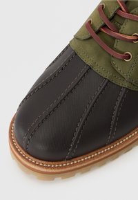 Chaussure en cuir noir texturé avec des accents en tissu vert, lacets marron et coutures crème. Bout rond avec une semelle en caoutchouc et des détails contrastés.