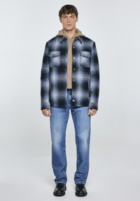 Camicia in flanella a quadri blu e nera con un hoodie sotto, abbinata a jeans azzurri chiari e stivali neri. Presenta due tasche sul petto.