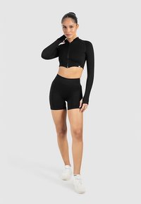 Černý crop top s dlouhými rukávy a zipem, kombinovaný s černými šortkami s vysokým pasem. Modelka má na sobě bílé tenisky; hladká látka, přiléhavý střih.