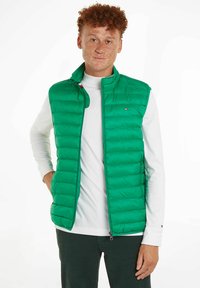 Grüne gepolsterte Weste mit hohem Kragen, ausgestattet mit einem Reißverschluss vorne, horizontalen Nähten und einem Logo-Detail in der Brusthöhe. Leichtes Design.