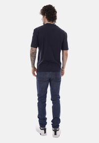 Uomo con capelli ricci di spalle, che indossa una maglietta nera, jeans blu, sneakers bianche e mostra tatuaggi sul braccio destro.