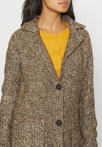 Stickad cardigan i brunt och beige med en strukturerad mönster, en stor krage och två mörka knappar, lager på lager över en gul button-up topp.