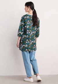Tunika w kwiatowy wzór w ciemnej zieleni, z białymi i niebieskimi kwiatami. Luźny krój, rękawy do 3/4, w połączeniu z jasno niebieskimi dżinsami i białymi sneakersami.