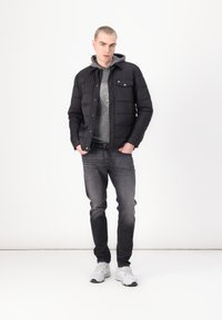 Chaqueta acolchada negra con una sudadera gris debajo, jeans grises y zapatillas gris claro. Presenta un bolsillo en el pecho y detalle del logotipo.