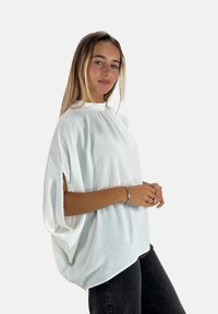 Maglietta bianca oversize con collo alto, maniche larghe con fessure laterali, realizzata in un tessuto leggero e leggermente testurizzato.