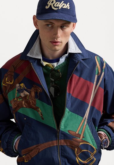 Veste en nylon colorée présentant des motifs équestres, avec des nuances de marine, vert, rouge et marron. Comprend une fermeture éclair et des poignets élastiques.