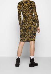 Svart långärmad klänning med ett gult paisley-mönster, figursydd design och knälång skärning, i kombination med svarta stövlar och vita strumpor.