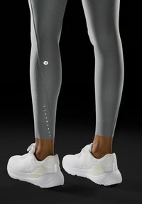 Szare legginsy z teksturowanym wzorem i odblaskowymi detalami; zestawione z białymi butami sportowymi z siateczkową cholewką i amortyzującą podeszwą.