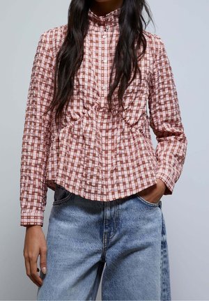 Femme portant un chemisier à carreaux roses et marron à boutons avec des détails bouffants, associé à un jean bleu clair ample, main dans la poche.