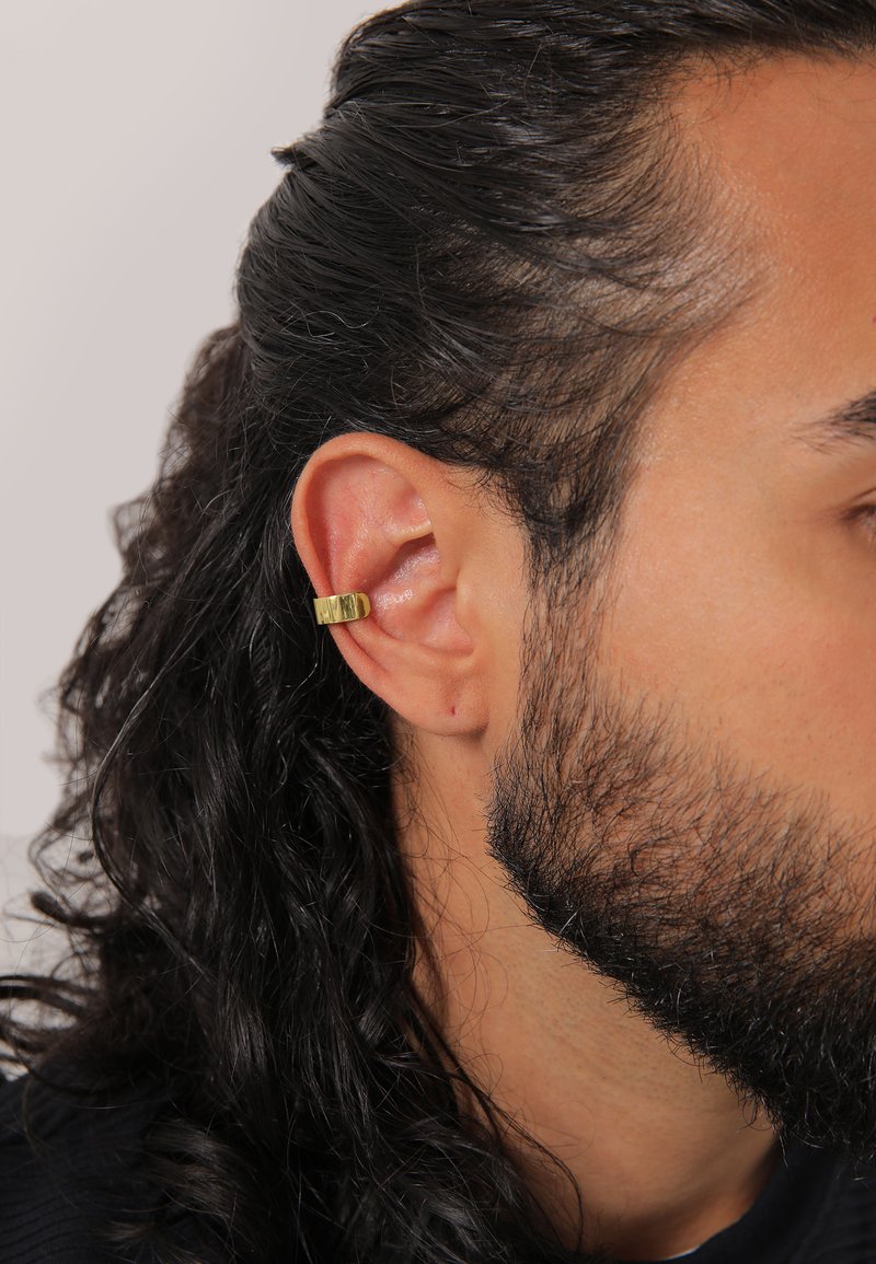 Guldfärgad ear cuff med en strukturerad yta, placerad på ett manligt öra med långt, mörkt, lockigt hår och en ljusbrun hy.