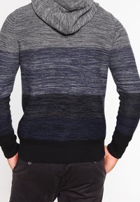 Man in een grijze, marineblauwe en zwarte gebreide hoodie met gradiëntstrepen, gezien van achteren, met de handen in de zakken.